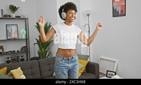 Fröhliche afroamerikanische Frau genießt Musik mit Kopfhörern in einem gemütlichen Wohnzimmer. Stockfoto