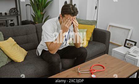 Ein gestresster junger Mann in Schutt, der zu Hause auf einer Couch sitzt und ein Stethoskop auf dem Tisch hat, was die Müdigkeit des Pflegepersonals darstellt. Stockfoto