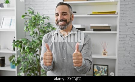 Hübscher hispanischer Seniorenmann mit grauem Bart, der die Daumen hoch in einer modernen Büroumgebung hält. Stockfoto