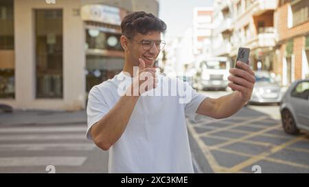 Junger hispanischer Mann, der ein Selfie auf einer städtischen Straße macht, während er einen Daumen nach oben zeigt, eine Brille und ein weißes T-Shirt trägt. Stockfoto