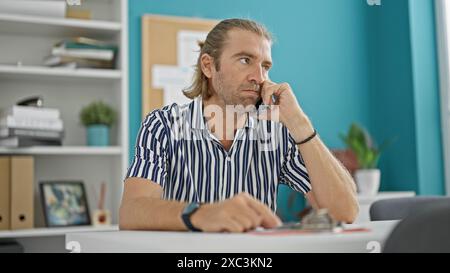 Hübscher langhaariger Mann in einem gestreiften Hemd, der an einem Telefon in einer modernen Büroumgebung spricht. Stockfoto