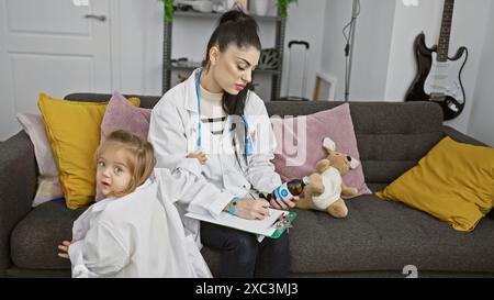 Eine fokussierte Frau in einem Labormantel, die auf ein Klemmbrett schreibt, während ein neugieriges Kleinkind in einem weißen Hemd neben ihr auf einer gemütlichen Couch in einem Zuhause sitzt. Stockfoto
