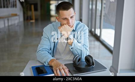 Ein nachdenklicher junger Mann, der in einer Universitätsbibliothek mit Laptop, Kopfhörern und Smartphone studiert. Stockfoto