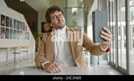 Hübscher hispanischer Mann mit Bart, der drinnen in einem modernen Büro ein Selfie macht, mit Brille und lässigem Rollkragen und Blazer. Stockfoto
