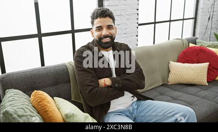 Hübscher junger Mann mit Bart, Arme kreuzen, sitzt bequem drinnen auf einer Couch in einem gut beleuchteten, gemütlichen Apartment. Stockfoto