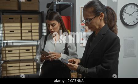 Zwei Frauen in einem Büro, eine untersucht ein Smartphone und die andere sieht sich Papiere an, was auf Ermittlungsarbeit hindeutet. Stockfoto