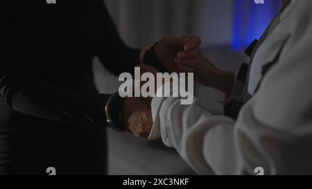 Eine Frau in einem weißen Blazer legt einem Mann hinter seinem Rücken Handschellen an, was auf eine Verhaftung in einem schwach beleuchteten Raum hindeutet. Stockfoto