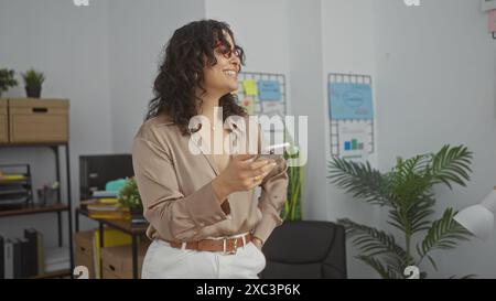 Junge hispanische Frau mit lockigen brünetten Haaren in einem Büro mit Brille und beiger Bluse, lächelnd, während sie ein Smartphone hält. Stockfoto