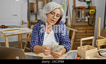 Eine reife Frau zählt polnische Zloty-Rechnungen in einer Tischlerei, während sie mit einem Smartphone spricht. Stockfoto