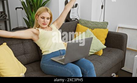 Blonde Frau, die sich auf einer grauen Couch mit Laptop in einem modernen Wohnzimmer entspannt Stockfoto