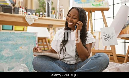 Eine lächelnde afroamerikanische Künstlerin, die ein Telefongespräch führt, während sie auf dem Boden in einem Studio sitzt. Stockfoto