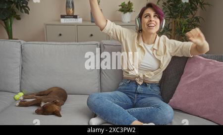 Junge Frau tanzt fröhlich auf einem Wohnzimmersofa mit einem Hund, der neben ihr schläft, und trägt Kopfhörer in einem gemütlichen Zuhause. Stockfoto