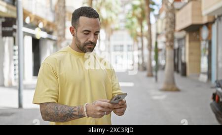 Ein gutaussehender hispanischer Mann mit Bart und Tätowierungen, der ein Smartphone auf einer hellen Straße benutzt Stockfoto