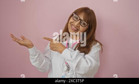 Eine schöne, reife, hispanische Frau mit einer Brille, die auf ihre ausgedehnte Hand zeigt, während sie vor einem isolierten rosa Hintergrund lächelt. Stockfoto