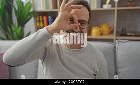 Ein lächelnder bärtiger Mann mittleren Alters macht die gute Geste über sein Auge drinnen, zu Hause. Stockfoto