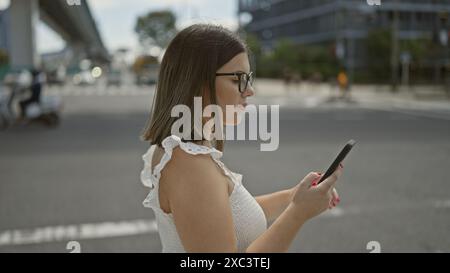 Wunderschöne hispanische Frau mit Brille, die selbstbewusst auf den Straßen tokios läuft, während sie in ihr Handy vertieft ist und eine ernsthafte geschäftliche SMS eingibt Stockfoto