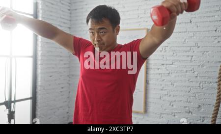 Junger chinesischer Mann, der Hanteln in einem Fitnessstudio hebt, sich intensiv auf sein Workout konzentriert und Stärke und Entschlossenheit zeigt. Stockfoto