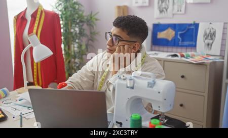 Ein nachdenklicher Mann mit Brille in einer modernen Schneiderwerkstatt, umgeben von einer Nähmaschine, Stoffproben und Designskizzen. Stockfoto
