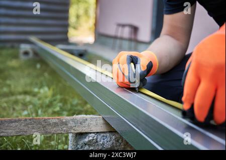 Nahaufnahme von Menschenhänden in Arbeitshandschuhen mit Bandmaß und Zeichenmarkierung auf Metallschiene mit Markierstift. Männlicher Arbeiter, der die Montageschiene aus Aluminium für die Installation von Solarpaneelen misst und markiert. Stockfoto