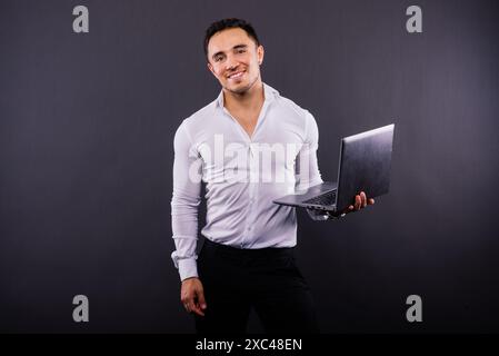 Bild eines jungen, fröhlichen Geschäftsmannes, der einen Laptop hält und benutzt, isoliert über dunklem Hintergrund Stockfoto