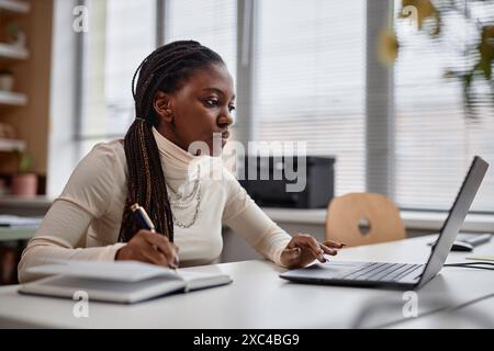 Porträt einer ernsthaften jungen afroamerikanischen Geschäftsfrau, die beim Schreiben in Notizblock auf Laptop-Computer recherchiert. Seitenansicht der durchdachten Schülerinnen, die sich auf die Prüfung vorbereiten, Kopierraum Stockfoto