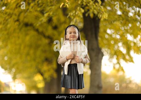 Kindertag. Fröhliches kleines Mädchen 5-6 Jahre alt posiert und lächelt draußen vor der Kamera und steht am Baum im Herbstpark. Stilvoller Zartpressdruck Stockfoto