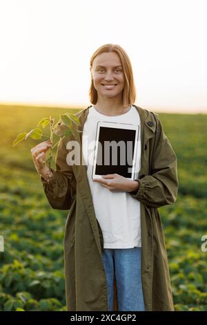 Die lächelnde Agronomin hält ein digitales Tablet mit Mockup-Bildschirm und Sojapflanze im Feld, untersucht, überprüft, schaut in die Kamera. Smart Farming so Stockfoto