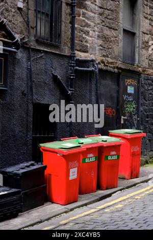 Rote, kommerzielle Recyclingbehälter aus Glas mit grünem Deckel, die vor einer schwarzen Wand außerhalb von angeordnet sind Stockfoto