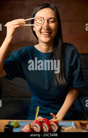 Positive asiatische Frau, die Sushi mit Stäbchen in der Nähe des Auges hält Stockfoto