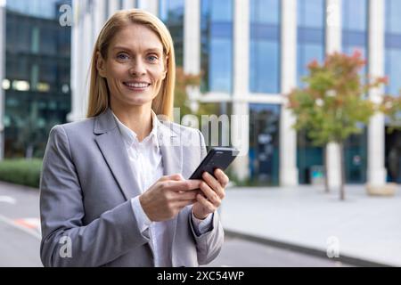 Nahaufnahme eines Porträts einer jungen lächelnden und erfolgreichen blonden Geschäftsfrau in einem Anzug, die vor einem Bürogebäude steht, ein Telefon hält und selbstbewusst in die Kamera schaut. Stockfoto