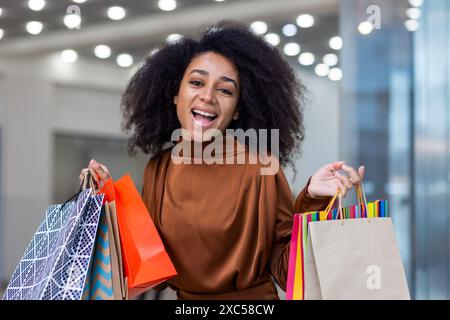 Porträt einer glücklichen jungen afroamerikanischen Frau, die mit Einkäufen und Rabatten zufrieden ist, eine Papiertüte mit Waren und Geschenken in der Hand hält und in die Kamera lächelt. Stockfoto