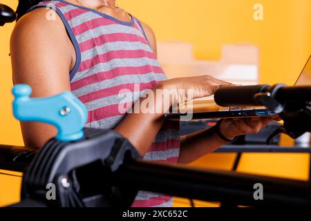 Nahaufnahme einer afroamerikanischen Person, die einen drahtlosen Computer für die Fahrradwartung hält. Das detaillierte Bild zeigt eine gesunde schwarze Frau, die einen Laptop neben dem Fahrradrahmen greift, der an einen Reparaturständer geklemmt ist. Stockfoto
