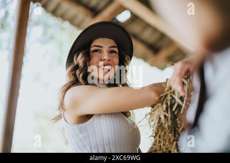 Eine junge Frau lächelt in der Scheune, trägt einen Hut und hält Heu Stockfoto