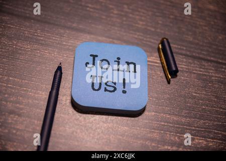 Das Konzept, das im Text „JOIN US“ auf einem Blatt vor einem mehrfarbigen Hintergrund vermittelt wird, deutet auf eine Einladung oder einen Aufruf hin, Teil von etwas zu sein. IT-Einflussnahme Stockfoto