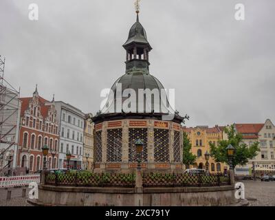 Historischer Brunnen auf einem Marktplatz, umgeben von alten Gebäuden und Kopfsteinpflasterstraßen unter bewölktem Himmel, Wismar, ostsee, deutschland Stockfoto