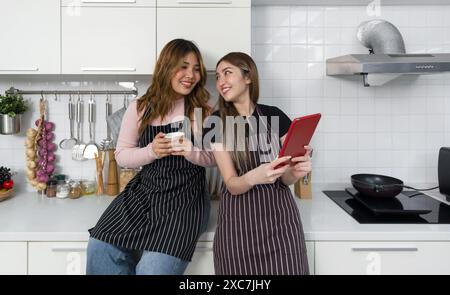 Zwei Frauen mit gestreifter Schürze, die in einer modernen Küche lesen oder mit einem Tablet-Computer interagieren. Kochutensilien und eine Auswahl an Dekorationen Stockfoto