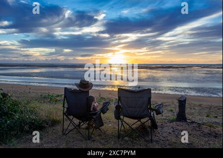 Männlicher Camper, der auf einem Stuhl am Strand sitzt und einen stimmungsvollen Sonnenaufgang beobachtet, Carmila Beach, Queensland, QLD, Australien Stockfoto