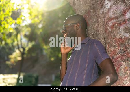 Ein junger afroamerikanischer Mann, der lächelnd und telefonierend am Telefon sitzt und sich an einem Baum im Freien lehnt - zwanglose und entspannte Unterhaltung - Natura Stockfoto