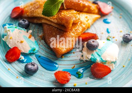 Pfannkuchen oder Crepe suzette mit Quark Creme, Erdbeeren, Heidelbeeren mit Blue Curacao Sauce und Orangenschale auf einem türkisfarbenen Teller. Nahaufnahme. Ausgewählter Fokus. Hochwertige Fotos. Hochwertige Fotos Stockfoto