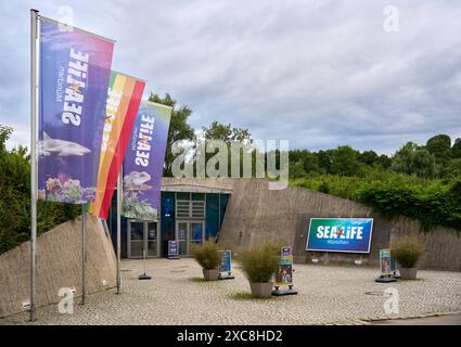 München, Bayern, Deutschland - 14. Juni 2024: Eingangsbereich von Sea LIFE München, einem beliebten Aquarium- und Freizeitpark in München, mit bunten Fahnen und Werbeschildern, umgeben von Natur und bewölktem Himmel *** Eingangsbereich des Sea LIFE München, einem beliebten Aquarium und Freizeitpark in München, mit farbenfrohen Flaggen und Werbeschildern, umgeben von Natur und einem bewölkten Himmel Stockfoto