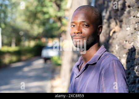 Ein junger afroamerikanischer Mann lächelt und lehnt sich an eine Wand und genießt einen sonnigen Tag im Freien - lässiges und entspanntes Porträt - natürlicher Hintergrund mit Stockfoto