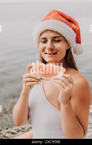 Eine Frau isst eine Wassermelone, während sie einen roten weihnachtsmann-Hut trägt. Die Szene ist unbeschwert und festlich, während die Frau einen erfrischenden Snack auf einem sunn genießt Stockfoto