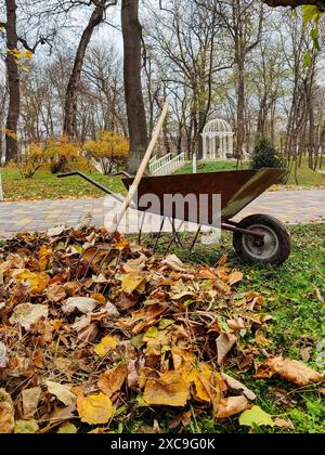 Rostige Schubkarre und Heugabel für die manuelle Ernte von in Haufen gesammelten Blättern im Herbstpark. Vor dem Hintergrund des weißen Pavillons. Warm, autonom Stockfoto