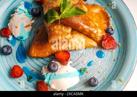 Pfannkuchen oder Crepe suzette mit Quark Creme, Erdbeeren, Heidelbeeren mit Blue Curacao Sauce und Orangenschale auf einem türkisfarbenen Teller. Nahaufnahme. Ausgewählter Fokus. Hochwertige Fotos. Hochwertige Fotos Stockfoto