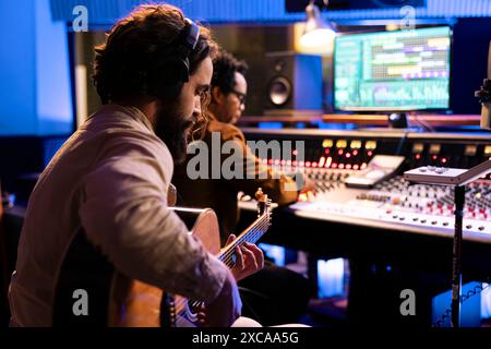 Songwriter kreiert einen neuen Song mit seiner Gitarre im Kontrollraum, spielt elektroakustische Instrumente und singt neben dem Plattenproduzenten. Junger Künstler, der live auftritt und Musik auf der Mischkonsole bearbeitet. Stockfoto