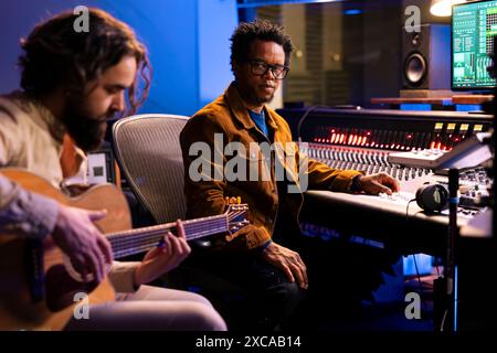 Der Sänger nimmt elektroakustische Gitarre in einem professionellen Musikstudio auf und spielt Musikinstrumente, um mit seinem Produzenten einen neuen Hit zu komponieren. Verschiedene Musiker singen eine Platte im Kontrollraum. Stockfoto