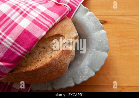 Ein frischer Brotlaib mit knusprigem französischem Brot, gewickelt in eine rot-weiße Serviette auf einem Metallteller auf einem hölzernen Bauernhof-Tisch Stockfoto