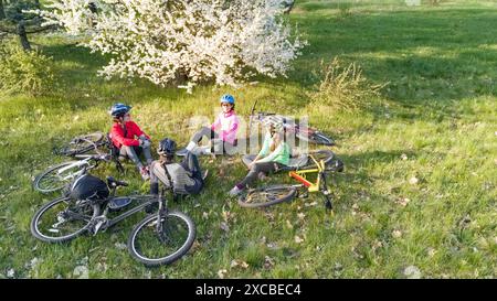 Familienradeln auf Fahrrädern im Frühling aus der Vogelperspektive von oben, fröhliche aktive Eltern mit Kindern haben Spaß und entspannen auf dem Gras unter blühenden Bäumen Stockfoto