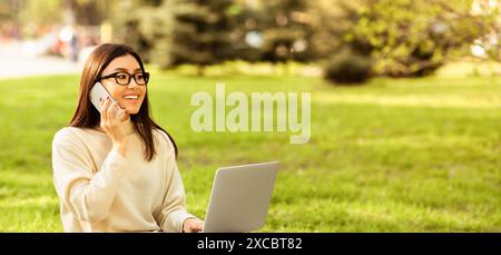 Frau sitzt im Gras mit Laptop und spricht am Telefon Stockfoto