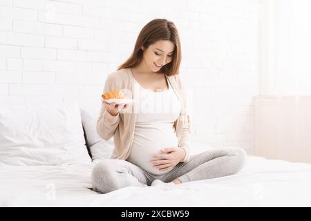 Schwangere Frau, die auf dem Bett sitzt und Croissant im sonnendurchfluteten Schlafzimmer isst Stockfoto